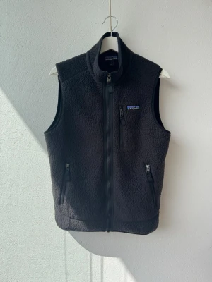 Svart fleeceväst från Patagonia -  Svart fleeceväst från Patagonia med hel dragkedja framtill. Västen har tre fickor med dragkedja och en liten logga på bröstet. Enkel att matcha med olika outfits. Nästan nyskick 9+/10