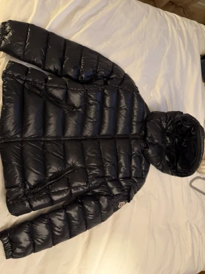Svart dunjacka från Moncler - Snygg svart dunjacka från Moncler med glansig finish, huva och två dragkedjefickor framtill. Jackan har en klassisk Moncler-logga på ärmen och quiltad design som håller värmen. Perfekt för kalla vinterdagar.