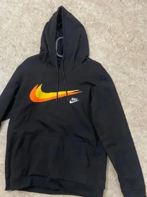 Svart Nike hoodie med färgglad logo - Svart hoodie från Nike med stor färgglad swoosh-logga i orange och gult på bröstet samt en mindre vit Nike-text. Klassisk känguruficka och dragsko i huvan. Perfekt för en sportig och avslappnad stil.