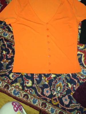 Orange topp med knappar och v-ringning - Snygg orange topp med korta ärmar och djup v-ringning. Toppen har knappar längs hela framsidan och är tillverkad i ett mjukt bomullstyg. Perfekt för dig som gillar färg och vill sticka ut lite extra.