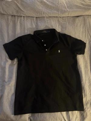 Svart pikétröja från Polo Ralph Lauren - Klassisk svart pikétröja från Polo Ralph Lauren med vit broderad logga på bröstet. Tröjan har korta ärmar, krage och knappar framtill. Perfekt för en stilren och avslappnad look.