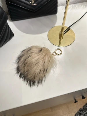 Hollies nyckelring  - Säljer en fluffig beige nyckelring med svarta toppar och guldfärgad metallring. Perfekt att hänga på väskan eller nyckelknippan för en trendig touch. Gjord i fuskpäls och metall. 