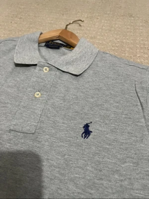 Polo Ralph Lauren pikétröja  - Klassisk grå pikétröja från Polo Ralph Lauren med korta ärmar och broderad marinblå logga på bröstet. Tröjan har krage, knäppning med två knappar och är tillverkad i mjuk bomull. Perfekt för en stilren och avslappnad look.