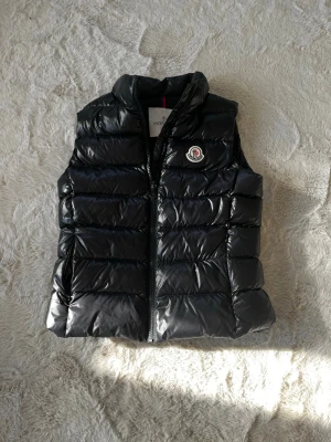 Svart dunväst från Moncler - Snygg svart dunväst från Moncler med glansig finish och klassisk Moncler-logga på bröstet. Västen har hög krage och dragkedja framtill. Perfekt för lager-på-lager och ger en clean streetwear-look.