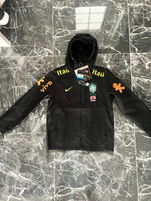 Svart Brasil Nike vindjacka med huva - Svart vindjacka från Nike med huva och dragkedja. Jackan har Brasilien landslagets emblem på bröstet, gula Nike-loggor och sponsortryck i gult och orange på ärmar och bröst. Perfekt för dig som gillar sportig stil och fotbollsinspirerade plagg.