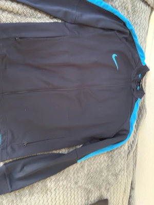 Nike svart träningsjacka XL - Svart träningsjacka från Nike med blå detaljer längs ärmarna och blå Nike-logga på bröstet. Jackan har hel dragkedja framtill, två fickor med dragkedja och är tillverkad i Dri-FIT-material som andas. Perfekt för träning eller sportaktiviteter.