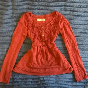 Lace babydoll top - Supersöt röd blus med långa ärmar, dekorativa knappar framtill och vacker spetsdetalj över bröstet. Blusen har en lätt utställd passform och markerad midja som ger en feminin siluett. Perfekt för dig som vill ha något extra till din outfit.