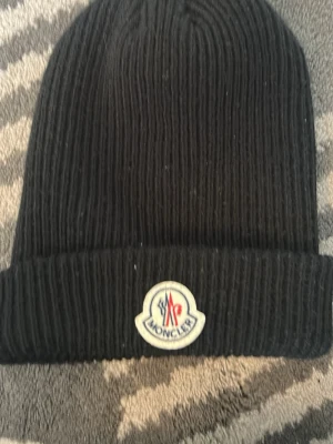 Svart ribbstickad mössa från Moncler - Säljer en svart ribbstickad mössa från Moncler med uppvikt kant och klassisk Moncler-logga framtill. Mössan är i mjukt material och har en enkel, stilren design som passar perfekt till kalla dagar.