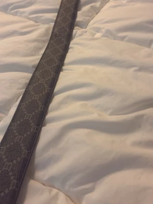 Svart B bälte med B-spänne - Snyggt svart bälte från B med mönstrad yta och ett stort B-format spänne. Bältet har ett diskret mönster med små B:n över hela bandet och är tillverkat i ett material som liknar skinn. Perfekt för att lyfta din outfit.