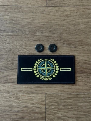 Stone Island 30 års patch - Säljer en svart patch från Stone Island med gul brodyr och gröna detaljer, samt två svarta knappar. Märket har en klassisk kompasslogga. Perfekt för att knäppa fast på jacka eller väska eller om du tappat bort din förra! Pris kan diskuteras!