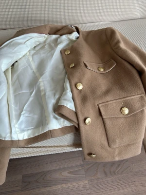 Beige kavaj med guldknappar - Stilren beige kavaj med fyra fickor framtill och snygga guldknappar. Kavajen har rak passform, vit insida och är tillverkad i ett ull-liknande material. Perfekt för dig som vill ha en klassisk och trendig look.