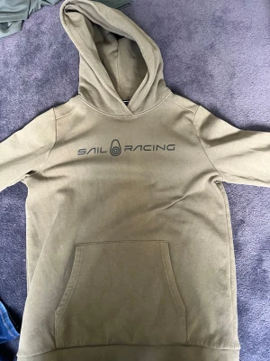 Olivgrön hoodie från Sail Racing - Olivgrön hoodie från Sail Racing med svart logga tryckt på bröstet. Klassisk känguruficka framtill och stor huva. Mjuk bomullskänsla och ribbade muddar vid ärmslut och nederkant. Perfekt för en avslappnad och sportig stil.