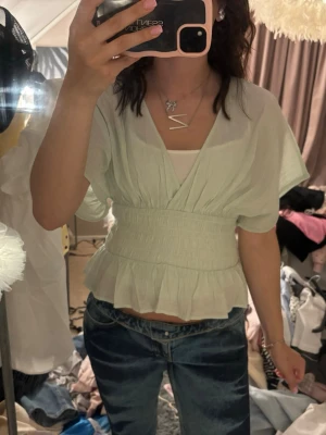 Ljusgrön blus med volang från Nelly - Supersöt ljusgrön blus från Nelly med korta ärmar, smockad midja och volang nertill. Djup omlottringning och lätt, luftig kvalitet som känns perfekt till jeans eller kjol. Materialet är tunt och ger en flowy känsla. Aldrig använd endast testad, stor i storleken så passar S bättre