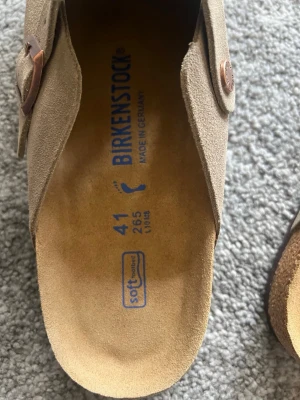Beige mockatofflor från Birkenstock - Säljer ett par snygga beige tofflor från Birkenstock i mjuk mocka. Modellen har en justerbar rem med metallspänne och präglad logga. Fotbädden är ergonomisk och sulan är i kork för extra komfort. Perfekta för chill dagar hemma eller ute. Storlek 41 låda medföljer ej 