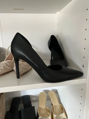 Svarta pumps från Boohoo - Stilrena svarta pumps från Boohoo med spetsig tå och hög smal klack. Skorna är tillverkade i ett slätt material som ger en elegant look. Perfekta för dig som vill ha en klassisk och tidlös sko till din outfit.