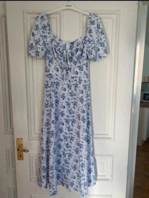 Blommig sommarklänning från SHEIN - Ljusblå och vit lång klänning från SHEIN med blommigt mönster. Klänningen har puffärmar, knytning framtill och en smickrande midja. Perfekt för varma dagar och har en romantisk vibe med sitt mjuka tyg och flowiga passform.