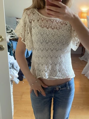 Vit spetsblus  - Söt vit blus i spets med korta, fladdriga ärmar och rund halsringning. Luftigt virkat mönster som ger en romantisk vibe. Perfekt till jeans eller kjol för en trendig look.