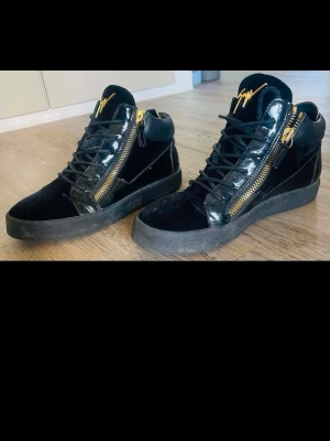 Svarta sneakers från Giuseppe Zanotti - Snygga svarta sneakers från Giuseppe Zanotti med guldfärgade dragkedjor och detaljer. Skorna har en mix av mocka och lackat läder, samt svart sula och snörning. Loggan syns i guld på plösen för en exklusiv vibe.