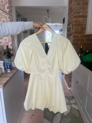 Ljusgul kort klänning med puffärmar - Superfin ljusgul kort klänning med stora puffärmar och djup v-ringning både fram och bak. Klänningen har knyt i ryggen och är tillverkad i ett lätt, slätt material som känns som bomull eller polyester. Perfekt för sommarens alla tillfällen!