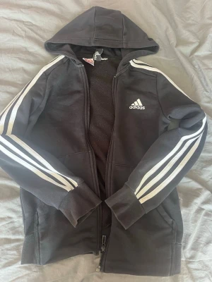 Svart Adidas hoodie med dragkedja - Svart hoodie från Adidas med klassiska vita ränder längs ärmarna och Adidas-logga på bröstet. Tröjan har dragkedja framtill, två fickor och en skön huva. Köp nu går bra eller skriv om du har frågor!🩷