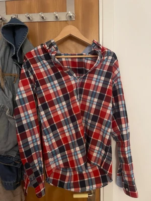 Rutig skjorta i blått och rött - Säljer en snygg rutig skjorta med klassisk krage och knappar framtill. Skjortan har en mix av blå, röd och vit färg och passar perfekt för dig som gillar en avslappnad och cool stil. Perfekt att bära över en t-shirt eller under en jacka.