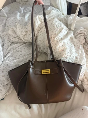 Brun handväska med guldspänne - Snygg brun handväska i slätt läder med långa axelband och ett stilrent guldspänne framtill. Väskan har en modern, rymlig design med utstående sidor och knytband. Perfekt för dig som gillar trendiga accessoarer.
