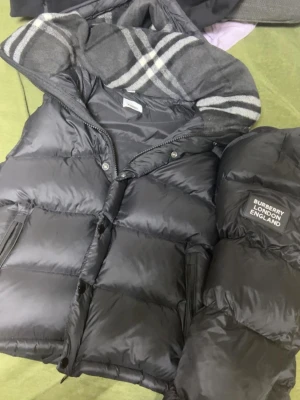 Burberry jacka med väst - Snygg svart dunväst från Burberry med klassiskt rutigt foder i huvan och dragkedja framtill. Västen har två fickor med dragkedja och en patch med 'Burberry London England' på bröstet. Perfekt för både sommar och vinter. Den passade inte därför jag säljer den. 