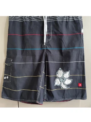 Svarta boardshorts med blommor  - Svarta boardshorts från Quiksilver med tunna färgglada ränder och en stor vit blomma som detalj. Shortsen har snörning i midjan och en praktisk ficka på sidan. Perfekta för stranden eller poolen.