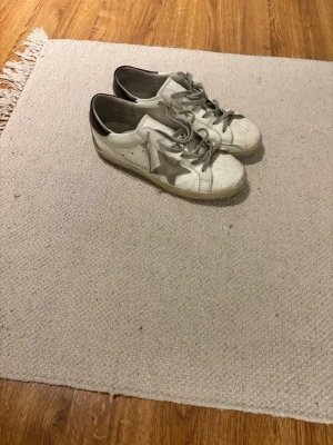 Golden Goose vita sneakers med stjärna - Snygga vita sneakers från Golden Goose med grå stjärna på sidan och grå skosnören. Skorna har svart detalj vid hälen och är tillverkade i läder. Klassisk låg modell med slitna detaljer för en cool look.