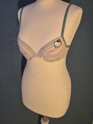 Hello Kitty randig bh med gröna band 75A - Söt bh från Hello Kitty med pastellfärgade ränder och gröna axelband. Dekorerad med Hello Kitty-tryck fram och text på banden. Uttagbara inlägg. Äkta Y2K. Aldrig använda, orginallappen från 2000-talet kvar. Perfekt för dig som gillar gulliga detaljer och färg. Justerbara axelband och klassisk knäppning bak. Se även mina andra varor med bundlerabatt. Finns också i storlek 75B, skriv till mig isf.