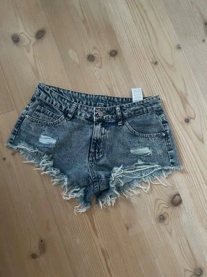 Grå slitna jeansshorts med fransar - Säljer ett par grå jeansshorts med medel midja, slitningar och fransig kant. Shortsen har fem fickor och bälteshällor. De är tillverkade i denim och har en cool, tvättad look med råa detaljer. Perfekta för en avslappnad och trendig stil. 