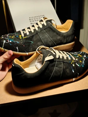 NFC ✅ Maison Margiela GATS svart sneakers paint  - NYPRIS ca7000kr, Mitt pris 2899kr. Maison Margiela sneakers i svart mocka och läder med unika färgstänk i vitt, gult och rött. Klassisk låg modell med vita snören och beige sula. Snygg och konstnärlig design som sticker ut. Kommer med originalkartong.