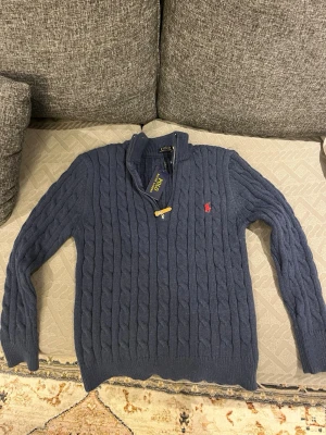 Blå kabelstickad tröja Polo Ralph Lauren - Helt ny mörkblå kabelstickad tröja från Polo Ralph Lauren med halv dragkedja och klassisk röd logga broderad på bröstet. Tröjan har ribbade muddar och krage, perfekt för en snygg och avslappnad stil.