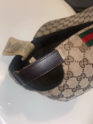 Gucci keps med GG-mönster - Snygg keps från Gucci i beige canvas med klassiskt GG-monogram över hela kepsen. Har justerbar rem i mörkbrunt läder och ikoniska gröna och röda ränder på sidan. Perfekt accessoar för att lyfta din streetwear-look.