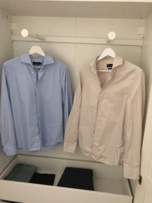 Beige & ljusblå kostymskjorta från Riley  - Kostymskjortor från Brothers av märke Riley i SLIM FIT. Ljusblå skjorta använd vid 2-3 tillfällen. Varsamt använd och tvättad. Inga skavanker. Beige skjorta använd 8-10 tillfällen. Varsamt använd och tvättad. Inga skavanker. Originalpris 700:- per skjorta. Köpt för knappt ett år sedan. Mitt pris är 300:- per skjorta och 500:- om man köper båda tillsammans. Storlek S - passar längd 175-185cm samt 65-75kg. 