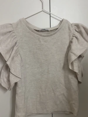Beige volangtopp från Zara - Supersöt beige topp från Zara med volangdetaljer längs ärmarna. Toppen har rund halsringning och är tillverkad i ett mjukt bomullsmaterial, sitter som xs/s 