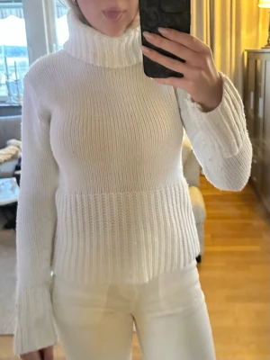 Vit stickad polotröja - Säljer en vit stickad polotröja med hög krage och långa ärmar. Tröjan har en ribbstickad struktur vid midjan och ärmsluten, vilket ger en snygg och modern passform. Perfekt för kyliga dagar och enkel att matcha med olika outfits.