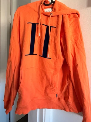 Orange hoodie från Les Deux - Snygg orange hoodie från Les Deux med stor svart II-logga på bröstet. Hoodien har huva med dragsko och en klassisk magficka framtill. Tillverkad i mjukt bomullsmaterial och har ribbade muddar vid ärmslut och nederkant. Perfekt för en avslappnad streetwear-look.