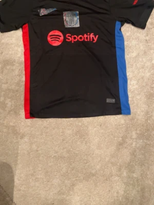 FC Barcelona svart matchtröja - Snygg svart matchtröja från FC Barcelona med röda och blå sidopaneler. Tröjan har Spotify-logga på bröstet och klubbens emblem i silver med röd och blå kant. Perfekt för fotbollsträning eller att bära som supporter. Materialet är lätt och andas bra.