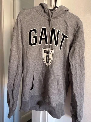 Grå hoodie från GANT med logga - Snygg grå hoodie från GANT med stor svartvit logga och emblem på bröstet. Klassisk känguruficka framtill, ribbade muddar och justerbar huva med dragsko. Perfekt för en chill och sportig stil.