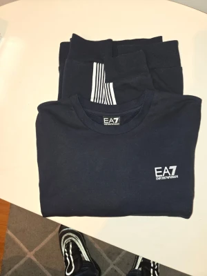Svart EA7 sweatshirt med vita ränder - Snygg svart sweatshirt från EA7 Emporio Armani med vita ränder längs ärmarna och logga på bröstet. Tröjan har rund halsringning och är tillverkad i mjukt material som passar perfekt till en sportig och stilren look. Som ny, storlek L passar M också.