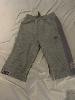 Grå mjukisbyxor från Reebok - Snygga grå mjukisbyxor från Reebok med bred resår i midjan och dragsko. Byxorna har en lila sidopanel med tryckt text och logga, samt patchdetalj vid benet. Perfekta för chill eller träning.