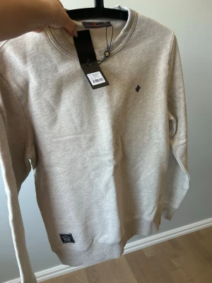 Beige sweatshirt från Morris - Ny - ej använd med etikett kvar. Snygg beige/khaki färgad sweatshirt från Morris med rund halsringning och diskret broderad logga på bröstet. Tröjan har ribbade muddar vid ärmslut och nederkant samt en liten patch nertill. Hör av dig om du har frågor eller undrar något. 