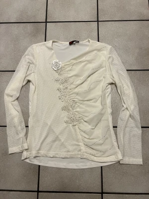 Vit långärmad topp med spets och pärlor storlek M/L - Vit långärmad topp med dekorativa spetsapplikationer och pärlor längs framsidan. Toppen har en lätt rynkad effekt och en söt blomdetalj vid axeln. Perfekt för dig som gillar unika detaljer och en feminin stil.
