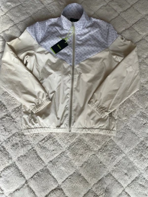 Beige och vit vindjacka från BOSS - Snygg beige vindjacka från Boss med vit panel upptill och diskret mönster. Jackan har dragkedja framtill, hög krage och elastiska muddar vid ärmar och nederkant. Perfekt för blåsiga dagar och enkel att matcha med olika outfits.