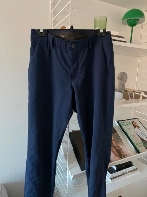 Mörkblå chinos från Under Armour - Snygga mörkblå chinos från Under Armour med klassisk rak passform och knappstängning i midjan. Byxorna har diskreta sidofickor och är tillverkade i ett lätt och bekvämt material som passar perfekt för en stilren look.