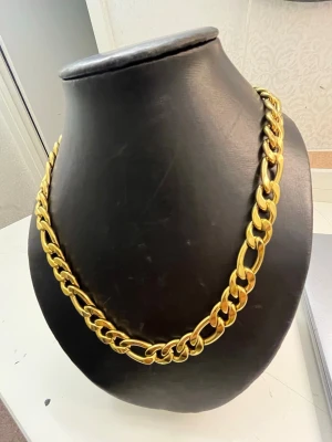 Kedjehalsband i guldtonad metall - Stilrent halsband med kraftiga, platta länkar i guldtonad metall. Kedjan har en klassisk design som ger ett statement-utseende och passar perfekt till att lyfta din outfit. Snygg och trendig accessoar för dig som gillar att sticka ut.
