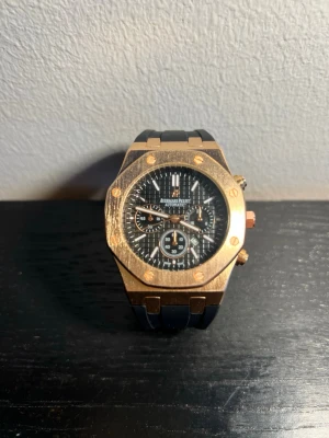 Audemars Piguet Royal Oak guld/svart - Snygg Audemars Piguet Royal Oak klocka med åttakantig boett i guldton och svart urtavla med flera subdials. Klockan har svarta detaljer och ett robust armband. Perfekt accessoar för dig som gillar exklusiv och modern stil. klockan är quartz och en av en, dem 2 knapparna och skruven fungerar inte, klockan är endast till för att ha på sig 