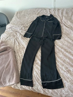 Svart satinpyjamas med vita detaljer - Snygg svart pyjamas  med vita kontrastsömmar längs kanterna. Setet består av en långärmad skjorta med krage och matchande byxor. Perfekt för mysiga kvällar hemma eller när du vill känna dig extra lyxig.