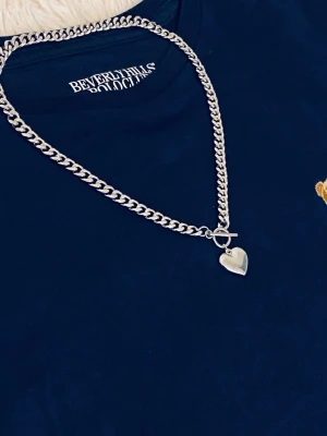 Silverfärgat kedjehalsband med hjärthänge - Stilrent halsband i silverfärgad rostfritt med kraftig kedja och ett hjärtformat hänge. Halsbandet har ett snyggt T-lås framtill som detalj. Perfekt accessoar för att lyfta din outfit med en cool och trendig touch.